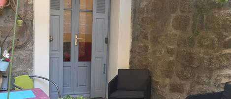 Een patio met een stenen muur, een deur met luiken en tuinmeubels, waaronder twee stoelen en een tafel.