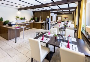 A modern hotel dining area with a kitchen counter, buffet, and several dining tables set with white chairs and pink napkins. - Montana Landhotel Gummersbach Nord (Marienheide)