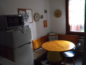 Cozinha compacta com geladeira, micro-ondas e mesa de madeira com cadeiras. Um relógio e um quadro decoram a parede.
