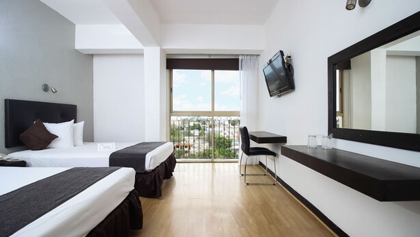 A hotel room with two beds, a desk, a chair, a TV, and a large window with a view. - Hotel Maya Yucatán (Mérida)
