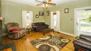 A living room with a brown leather sofa, a brown armchair, a coffee table, and a patterned rug. There is a ceiling fan, a fireplace, and a door leading outside. - 2 Bedroom 1 Bath Cottage (Knoxville)