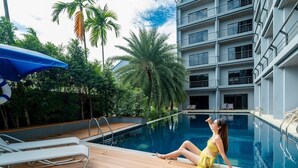 A poolside area with lounge chairs, a palm tree, and a modern building in the background. - Bhukitta Boutique Hotel - SHA Plus (PHUKET)