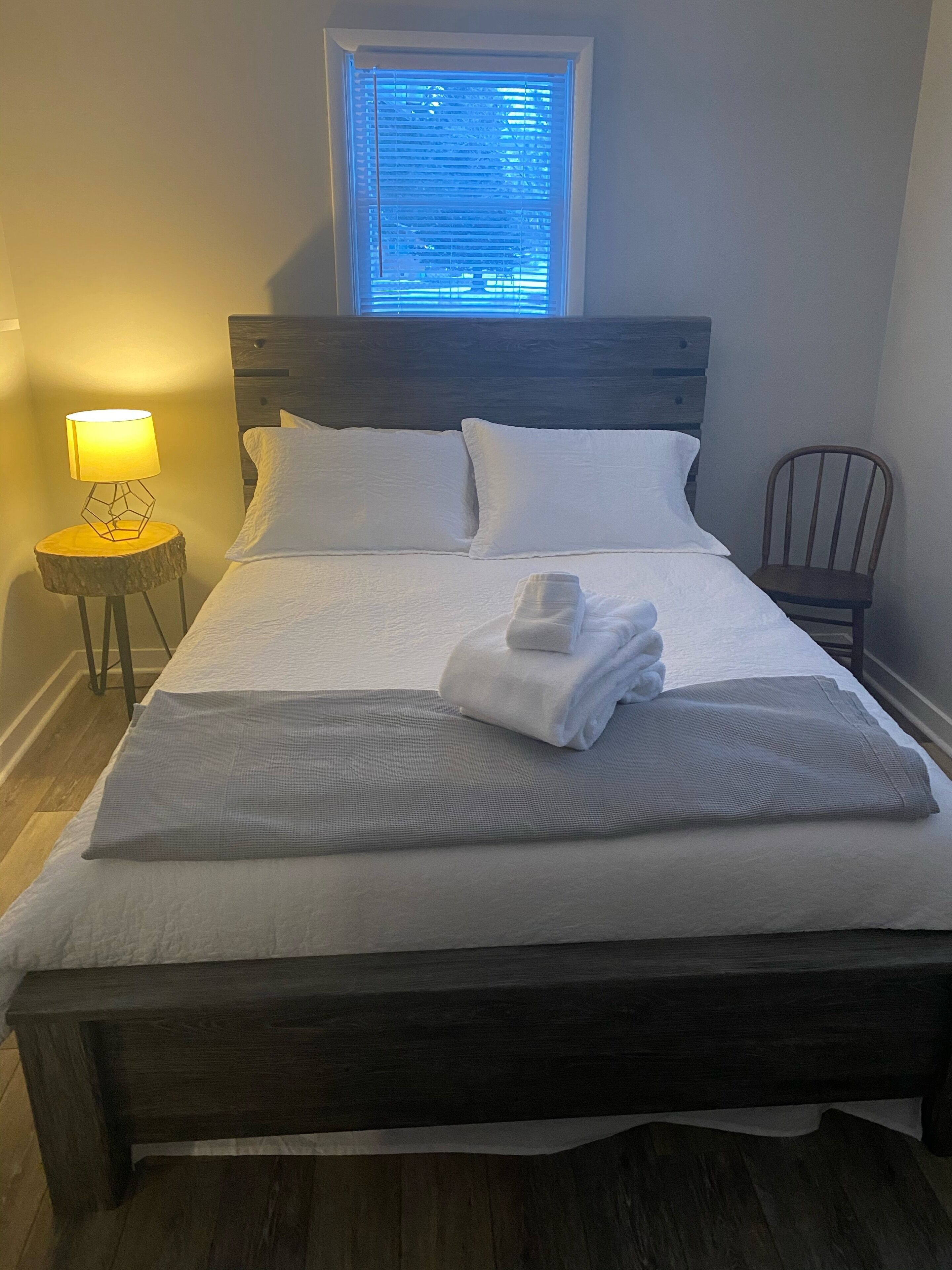 A neatly made bed with white linens, a wooden headboard, and a window with blinds.