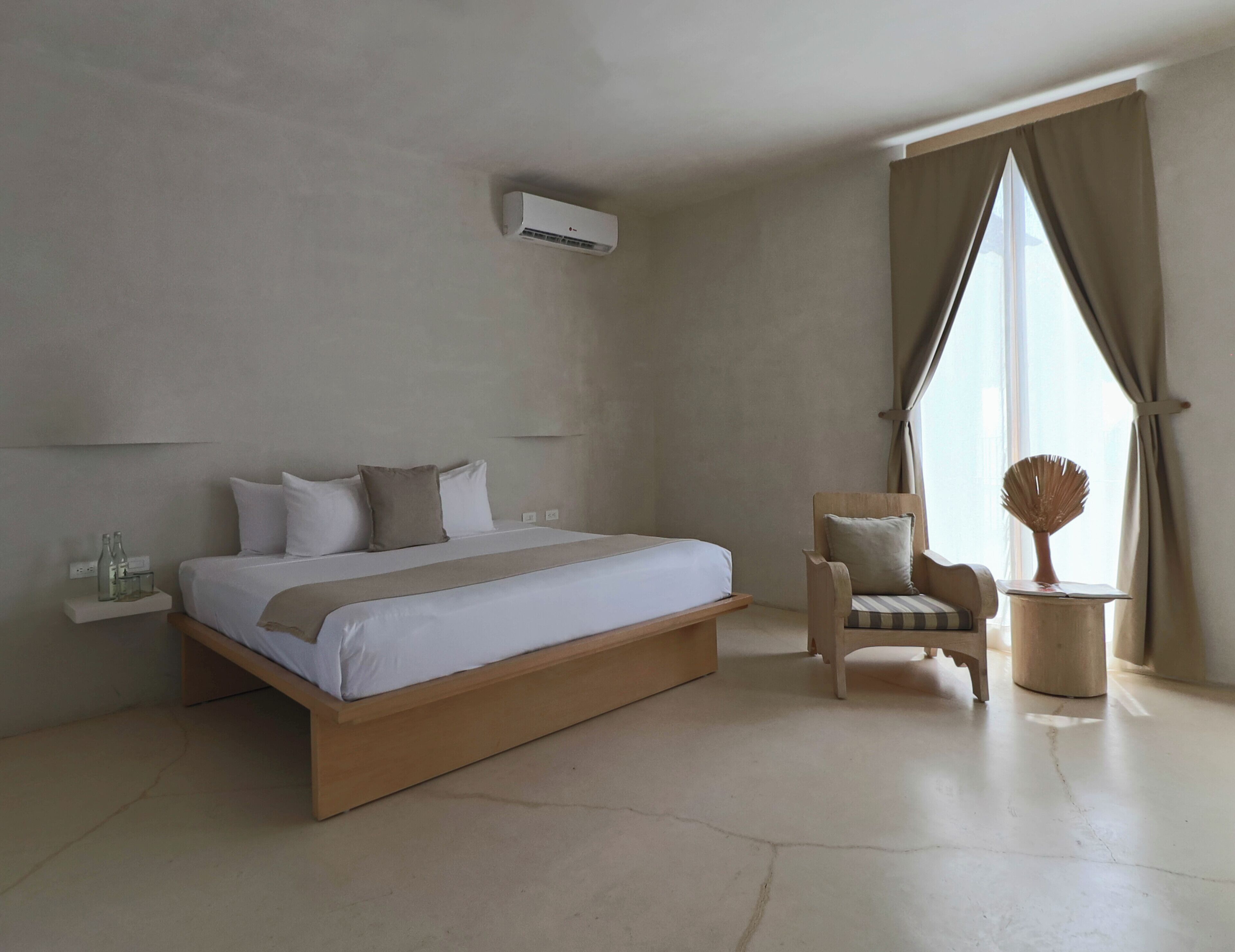A modern hotel room with a large bed, a chair, a small table, and a window with curtains.