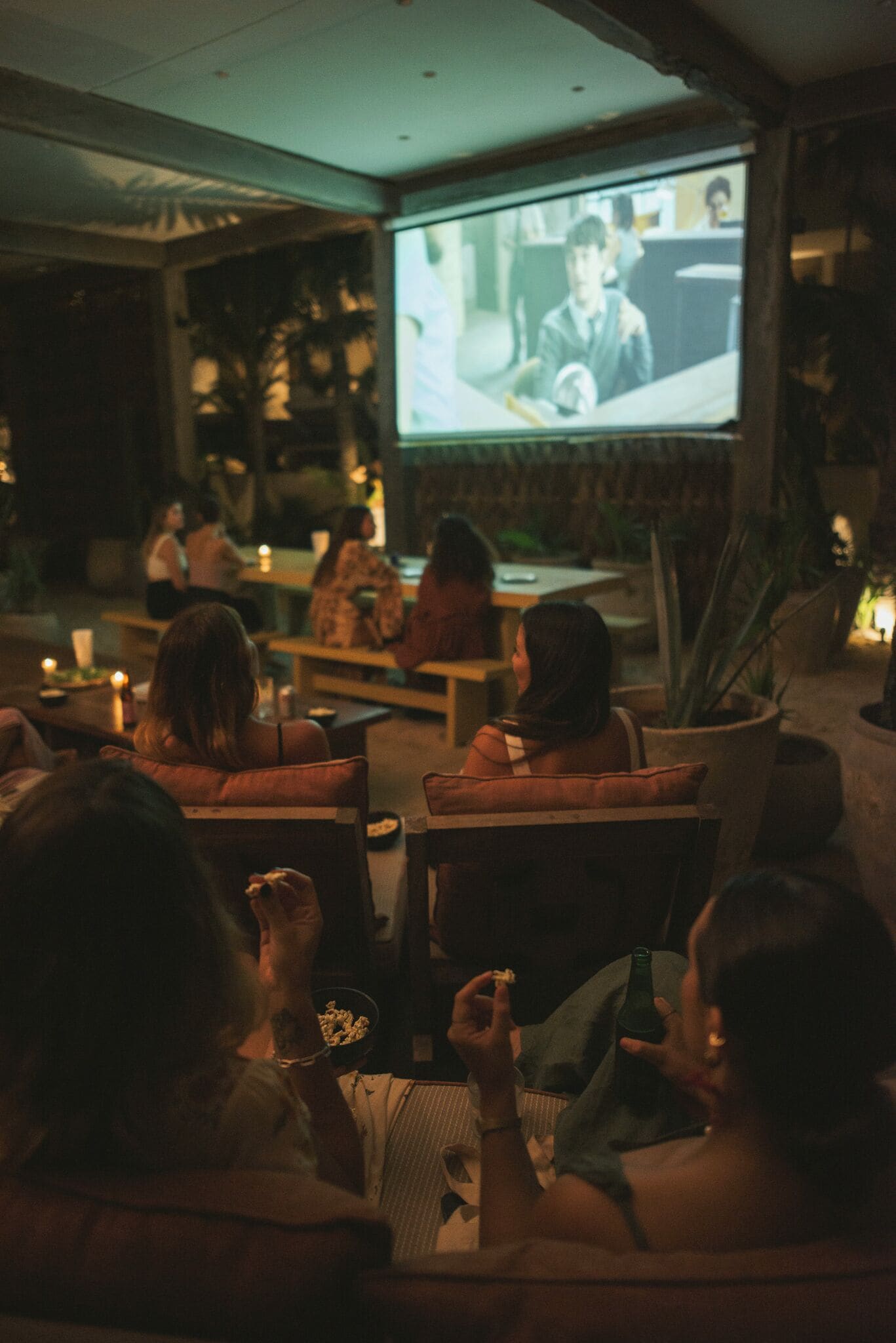 Una habitación tenuemente iluminada con un grupo de personas sentadas en sofás viendo una película en una pantalla grande.