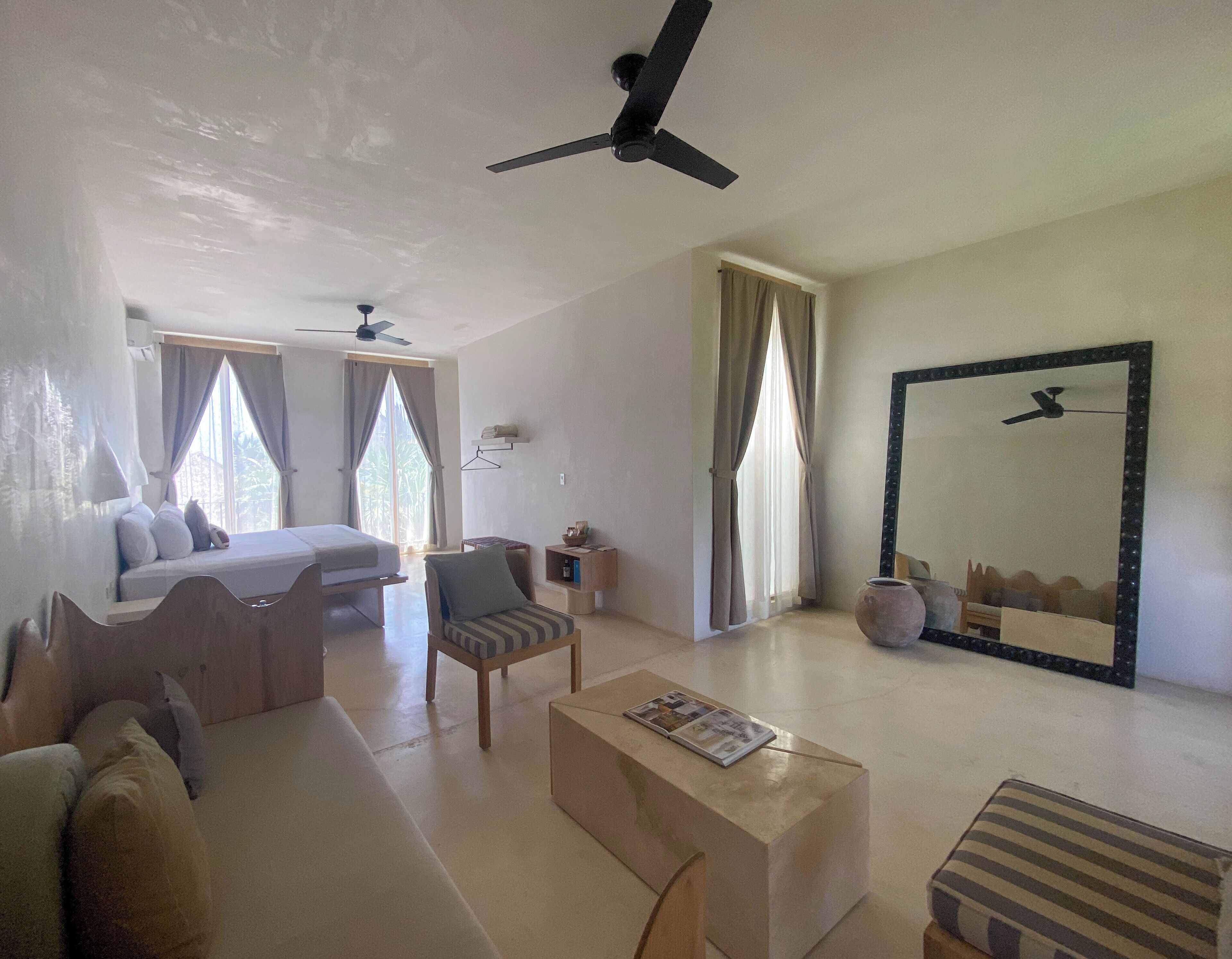 A hotel room with a bed, a sofa, a chair, a small table, and a large mirror. There are ceiling fans and a window with curtains.