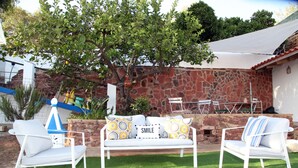 Un espace patio aménagé avec un ensemble de meubles de jardin blancs, un mur en pierre et un arbre qui offre de l’ombre.