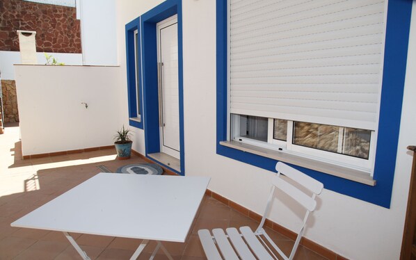 A patio with a white table and a white chair, a potted plant, and a window with blinds.
