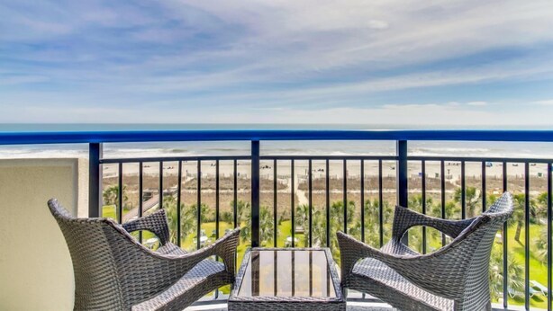 Een balkon met rieten meubels en een glazen tafel, met uitzicht op een strand.