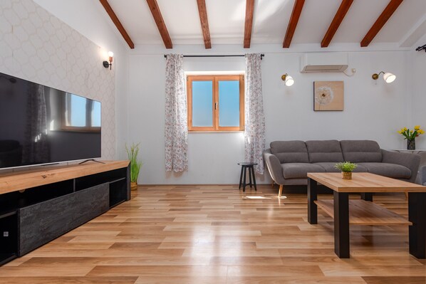 Un salon doté d’un téléviseur à écran plat, d’un parquet, d’un canapé gris, d’une table basse en bois et d’une fenêtre ornée de rideaux à motifs floraux.