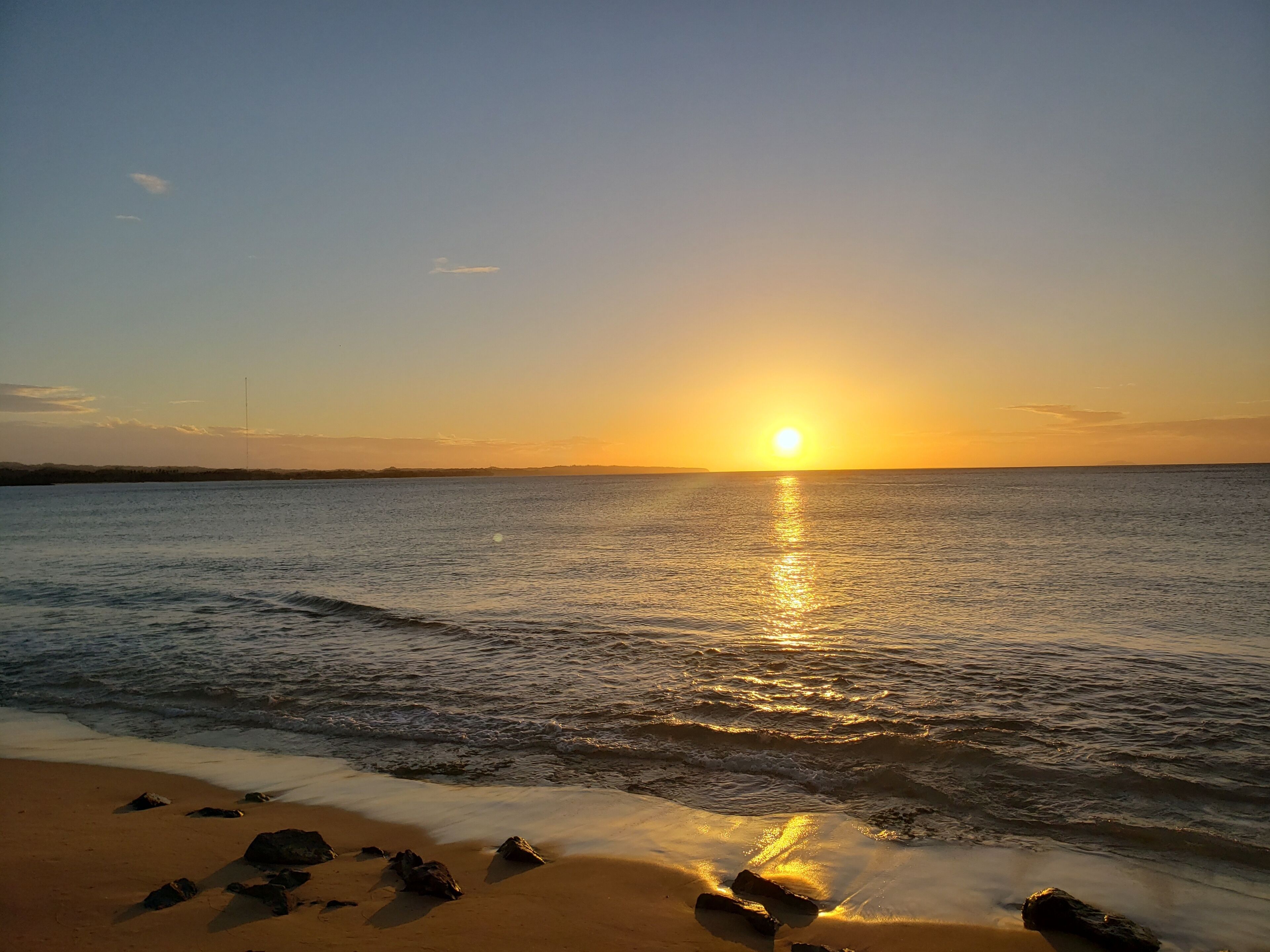 A beach with the sun setting over the horizon, calm sea, and scattered rocks on the shore.