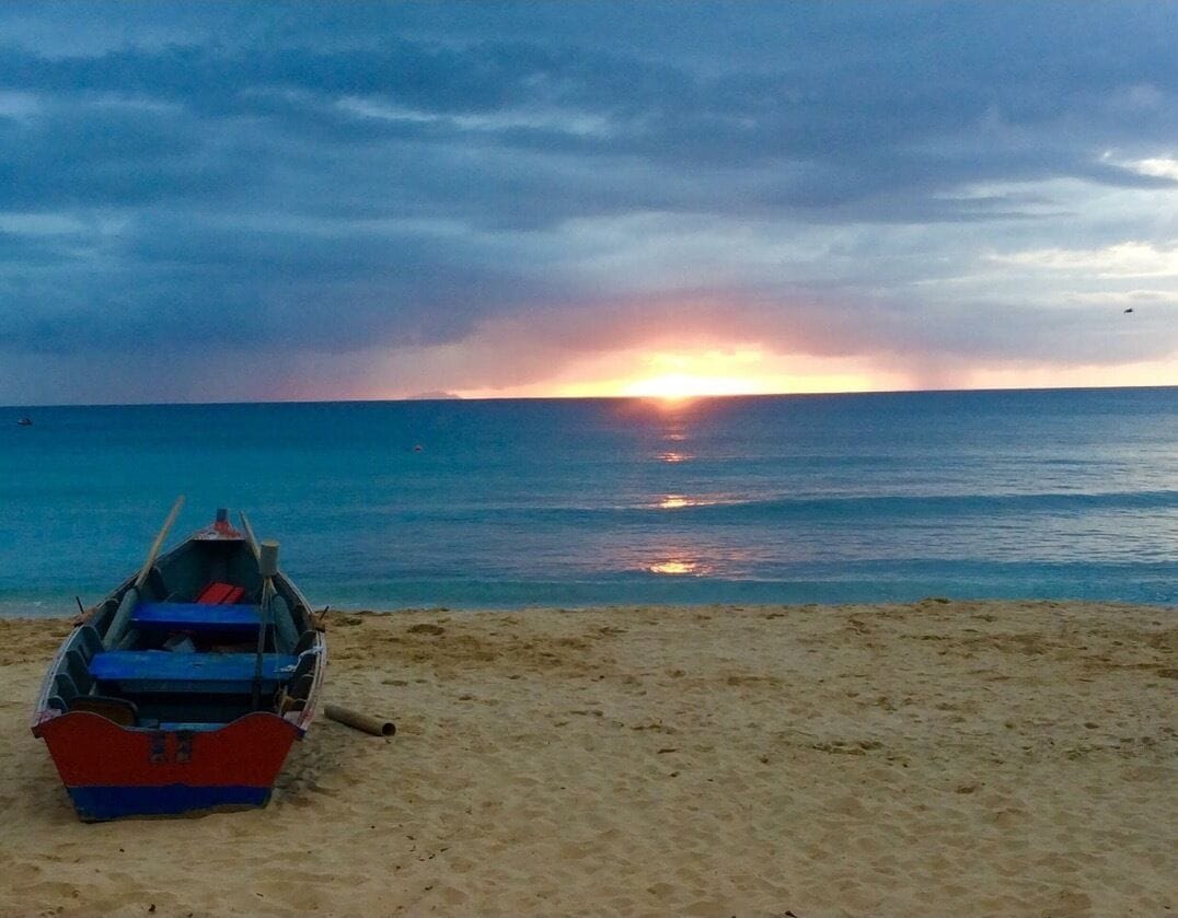 A boat on a sandy beach with a sunset over the ocean.