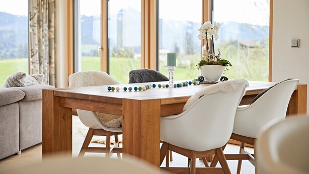A dining area with a wooden table, white chairs, and a view of greenery through large windows.