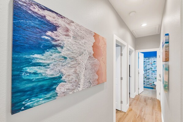 A hallway with a large abstract painting of a wave on the wall, white doors, and a view into another room with patterned wallpaper.