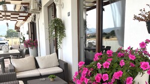 Uma área de estar externa com móveis de vime, um grande vaso com flores cor-de-rosa e vista para a paisagem através de uma porta de vidro.