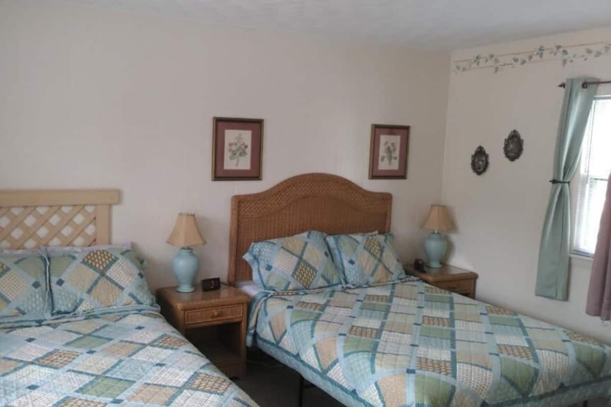A bedroom with two beds, each with a headboard and nightstands. There are framed pictures on the wall and a window with curtains.