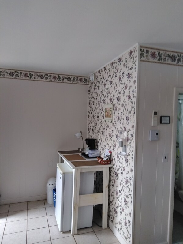 A small, clean room with a desk, a coffee machine, a trash bin, and a door leading to another room. - Midway Lodging (Cornish)