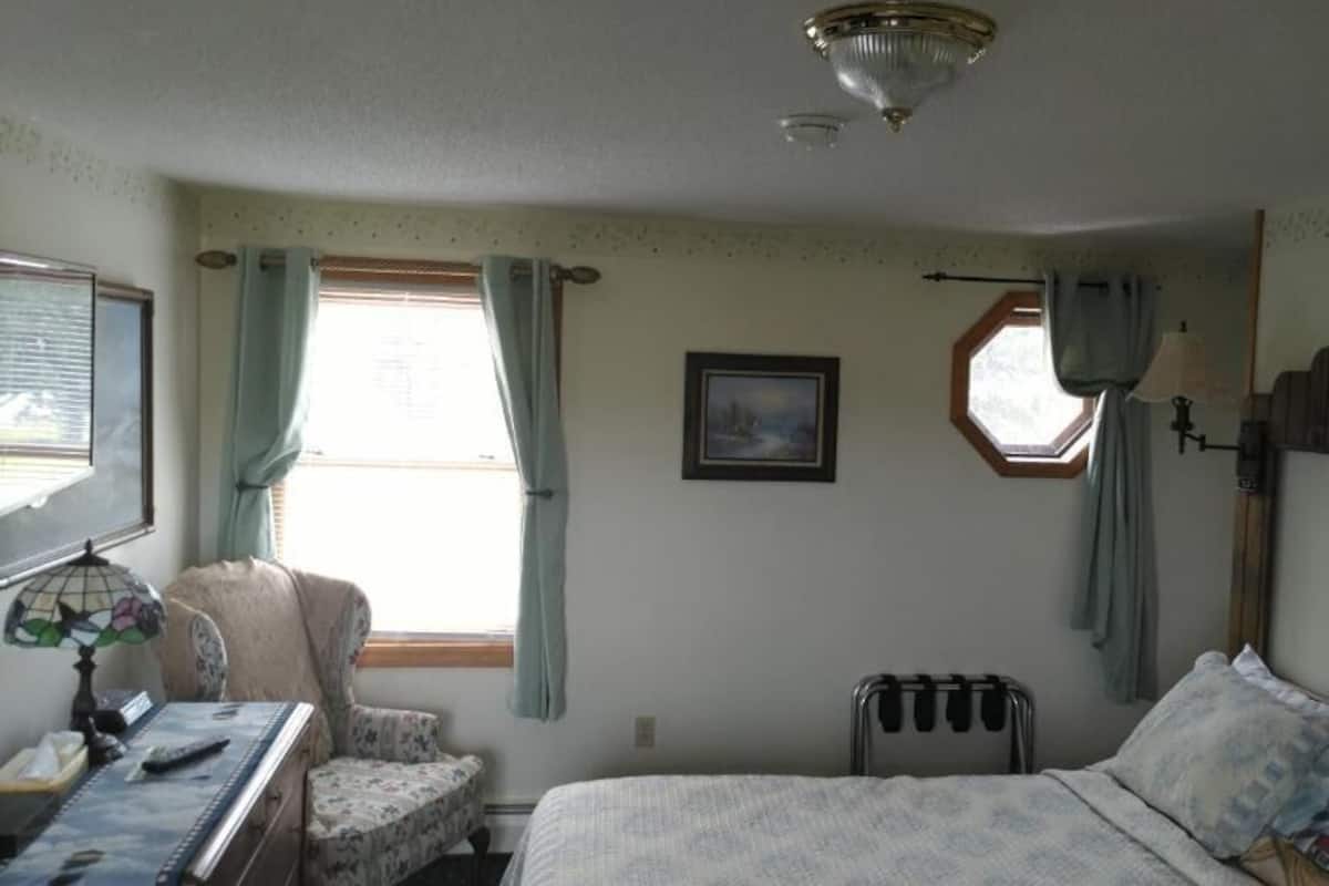 A bedroom with a bed, a desk with a lamp, a chair, a radiator, and a window with curtains.