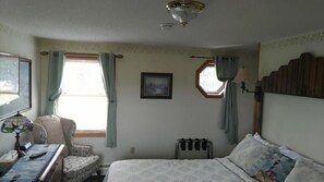 A bedroom with a bed, a desk with a lamp, a chair, a radiator, and a window with curtains. - Midway Lodging (Cornish)