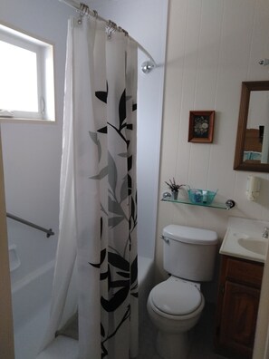 A bathroom with a toilet, a glass shelf with toiletries, a framed picture on the wall, and a shower curtain with a leaf pattern. - Midway Lodging (Cornish)