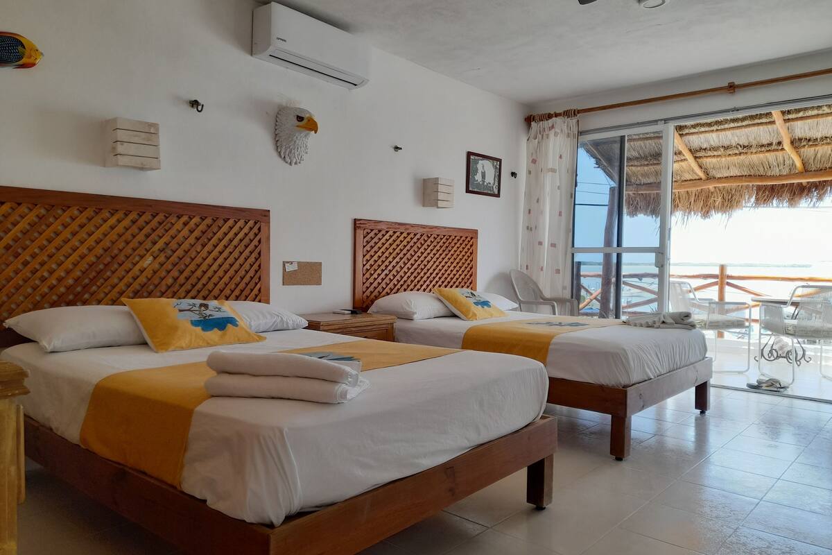 A hotel room with two wooden beds, a ceiling fan, an air conditioning unit, and a view of the beach through large windows.