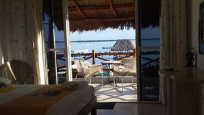 A room with a bed, a table, and a view of a beach with boats and a thatched-roof structure. - Hotel Punta Ponto (Río Lagartos)