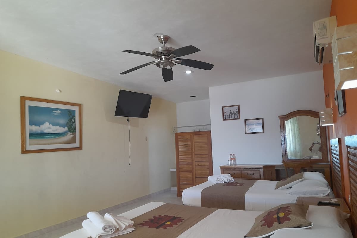 A hotel room with two beds, a ceiling fan, a flat-screen TV, a painting of a beach scene, and a wooden wardrobe.
