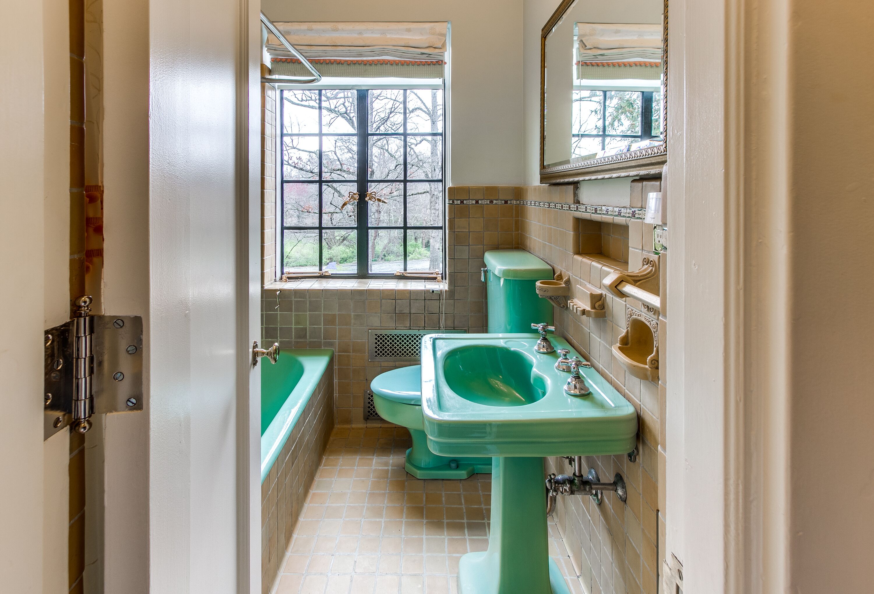 Une salle de bain avec un lavabo, des toilettes et une baignoire, le tout de couleur verte. Une fenêtre offre une vue sur les arbres à l’extérieur.