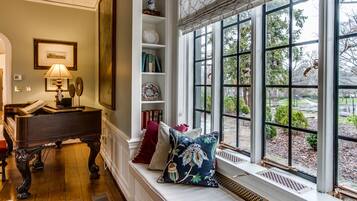 A room with a piano, a bench with cushions, a bookshelf, and a window with a view of greenery.