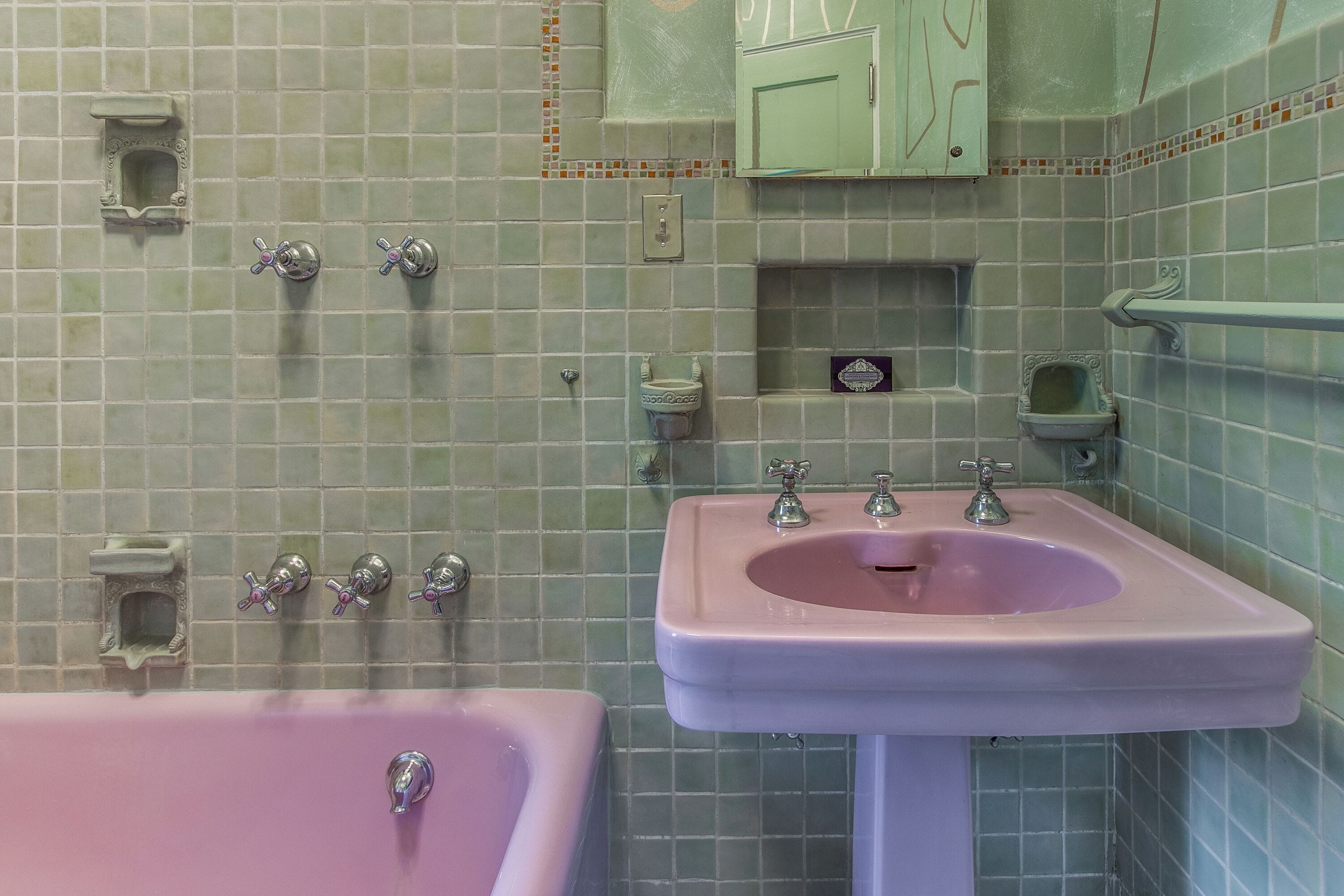 Een badkamer met een roze badkuip, een wastafel met twee kraan en een groen kastje boven de wastafel.