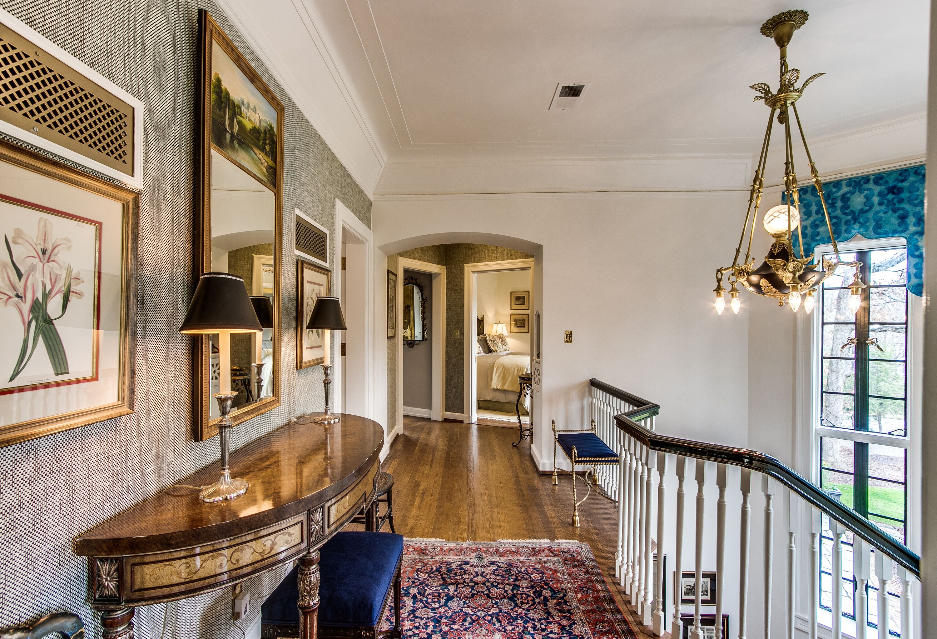 A hallway with a wooden floor, a blue patterned rug, a chandelier, and a staircase with white railings.