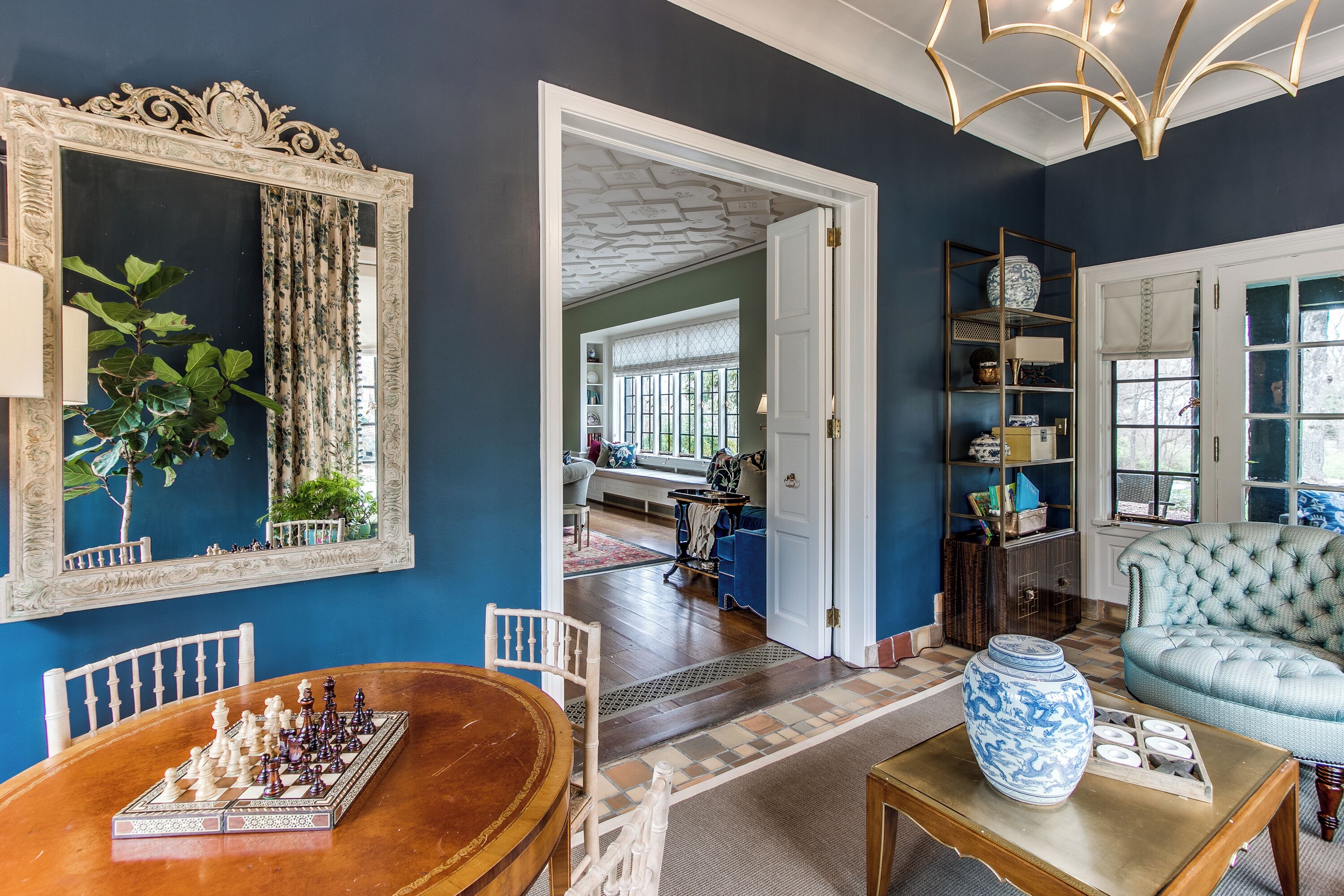A room with a chessboard, a large mirror, a blue armchair, and a vase on a table.