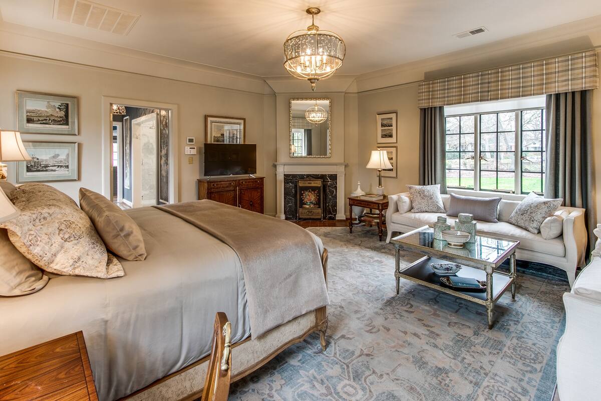 A spacious bedroom with a large bed, a sitting area with a sofa, a fireplace, and a television.