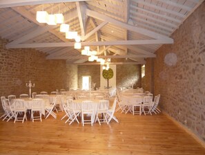 A spacious hall with high ceilings and exposed beams, furnished with round tables and white chairs, suitable for events. - Homerez - Manor in Satillieu (Satillieu)