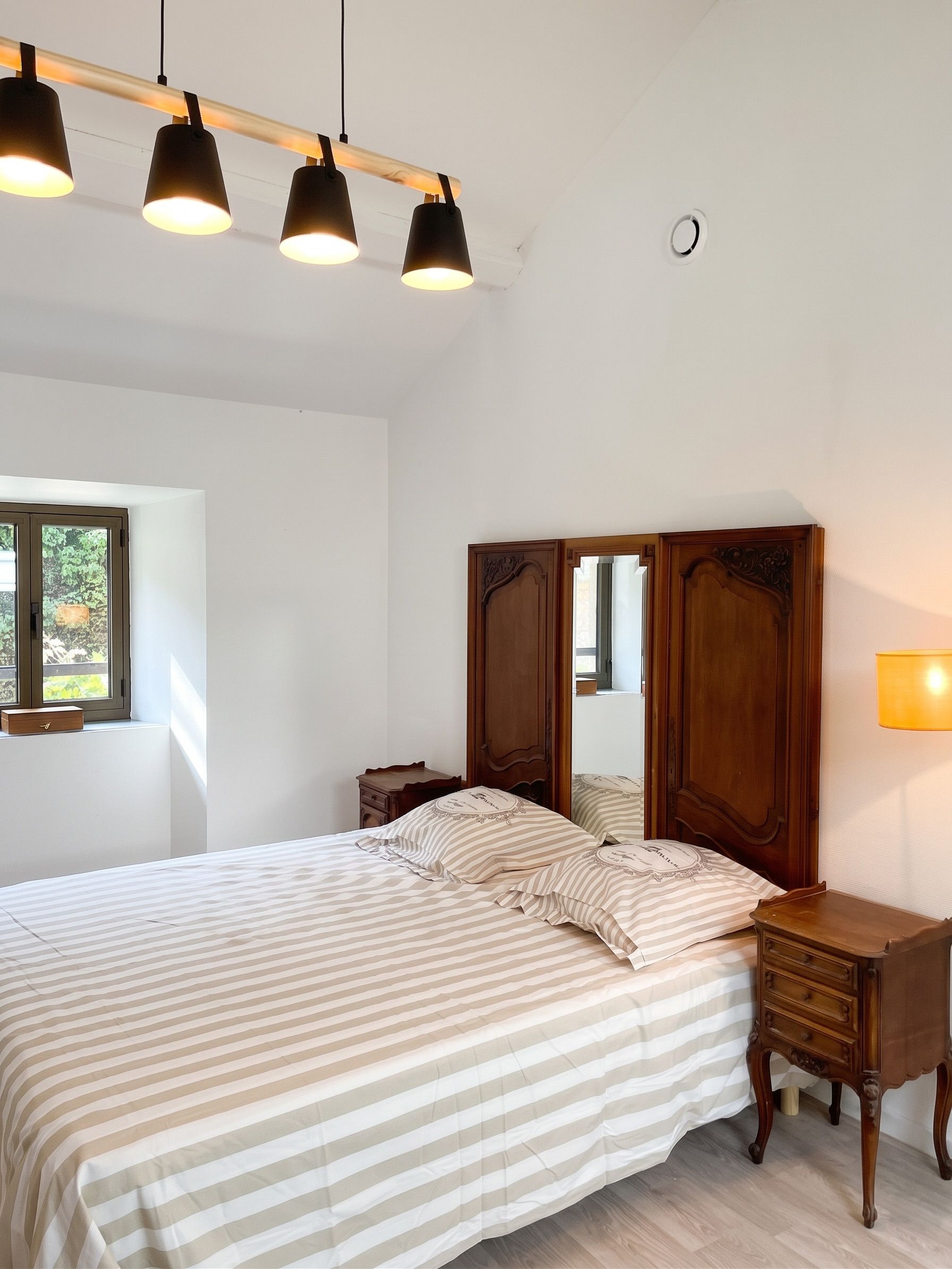 A bedroom with a bed, bedside tables, a wooden headboard, a window, and a wall-mounted lamp.