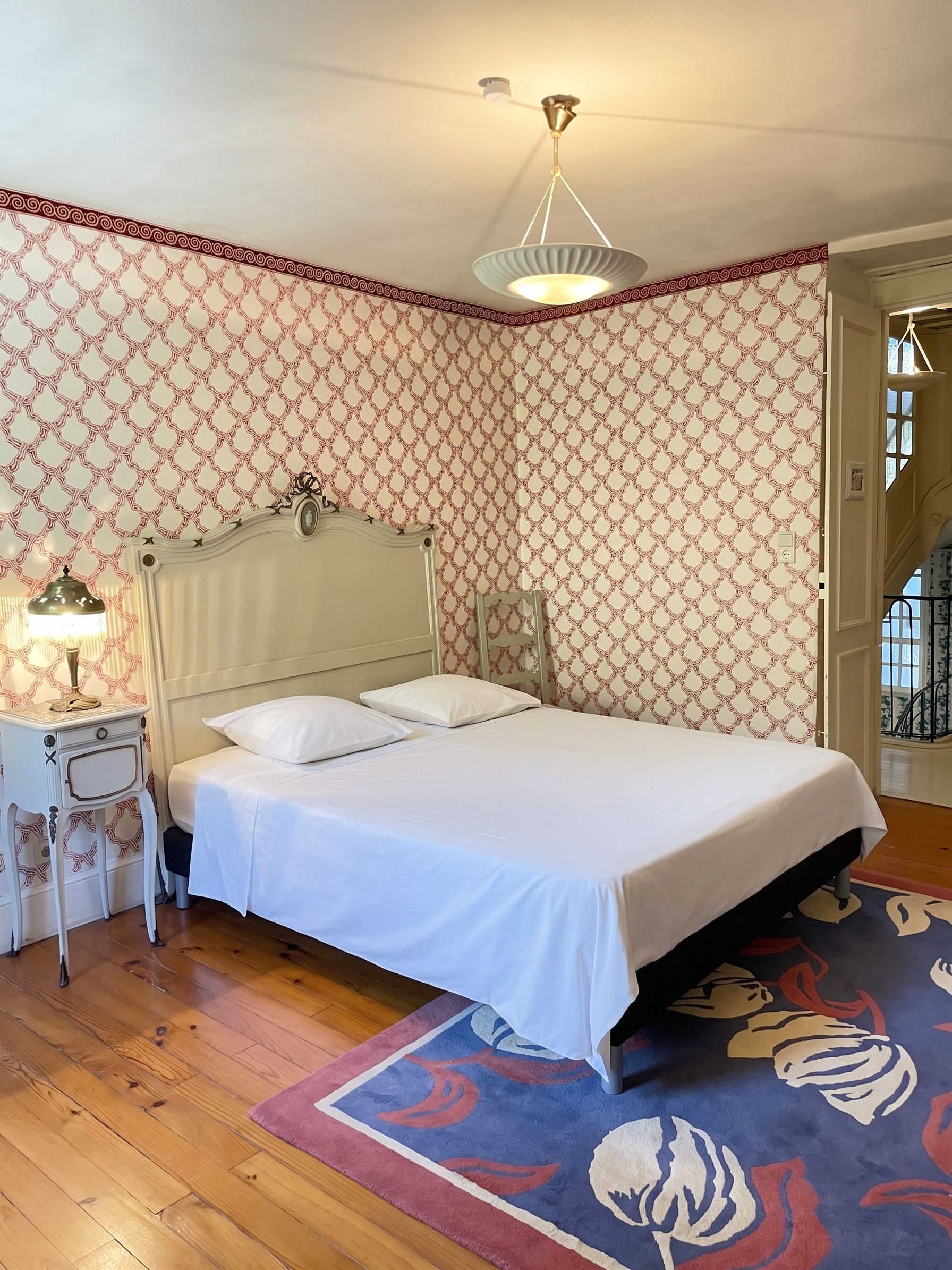 Un dormitorio con cama, mesita de noche, lámpara, papel tapiz estampado y suelo de madera.