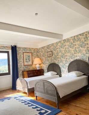 A bedroom with two single beds, a window with a view, a wooden dresser, and a blue patterned rug. - Homerez - Manor in Satillieu (Satillieu)