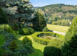 A landscaped garden with various trees and shrubs, a fenced area, and rolling hills in the background. - Homerez - Manor in Satillieu (Satillieu)