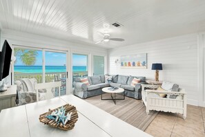 A spacious living room with a large window overlooking the ocean, a white ceiling fan, and a cozy seating area with a white crib.