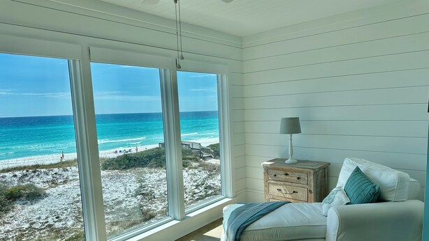Una habitación con un ventanal grande que da a una playa, una silla blanca con cojín, una mesita de noche de madera con lámpara y un ventilador de techo.