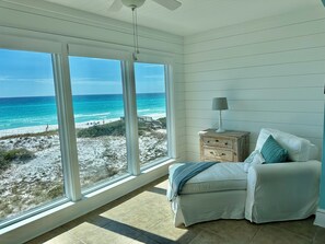 A room with a large window overlooking a beach, a white chair with a cushion, a small wooden nightstand with a lamp, and a ceiling fan. - Community Beach Access, Adorable Gulf Front Condo, 4BR, pool/hot tub (Santa Rosa Beach)
