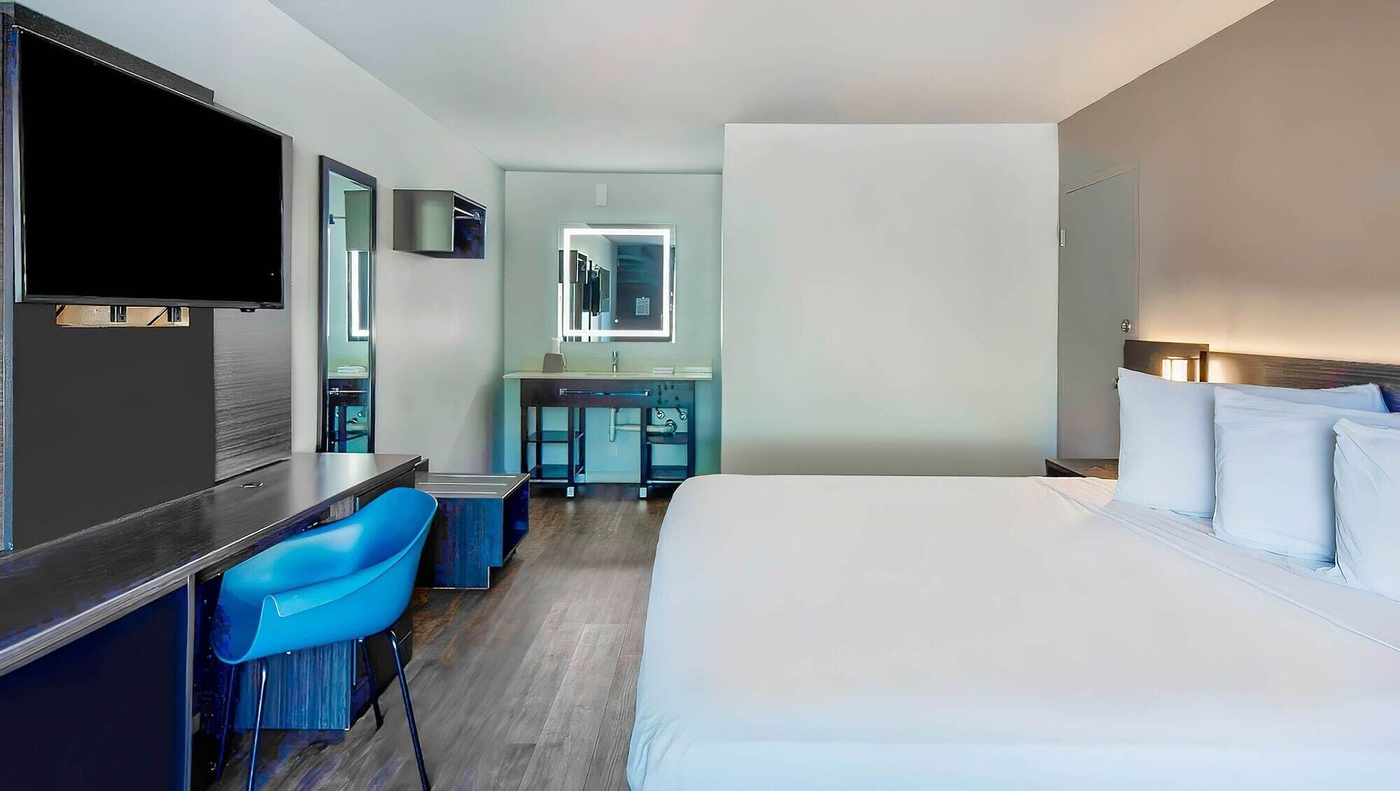 Ein Hotelzimmer mit einem Bett, einem Schreibtisch mit Stuhl, einem Fernseher, einem Spiegel und einem Badezimmer mit Waschbecken und Spiegel.
