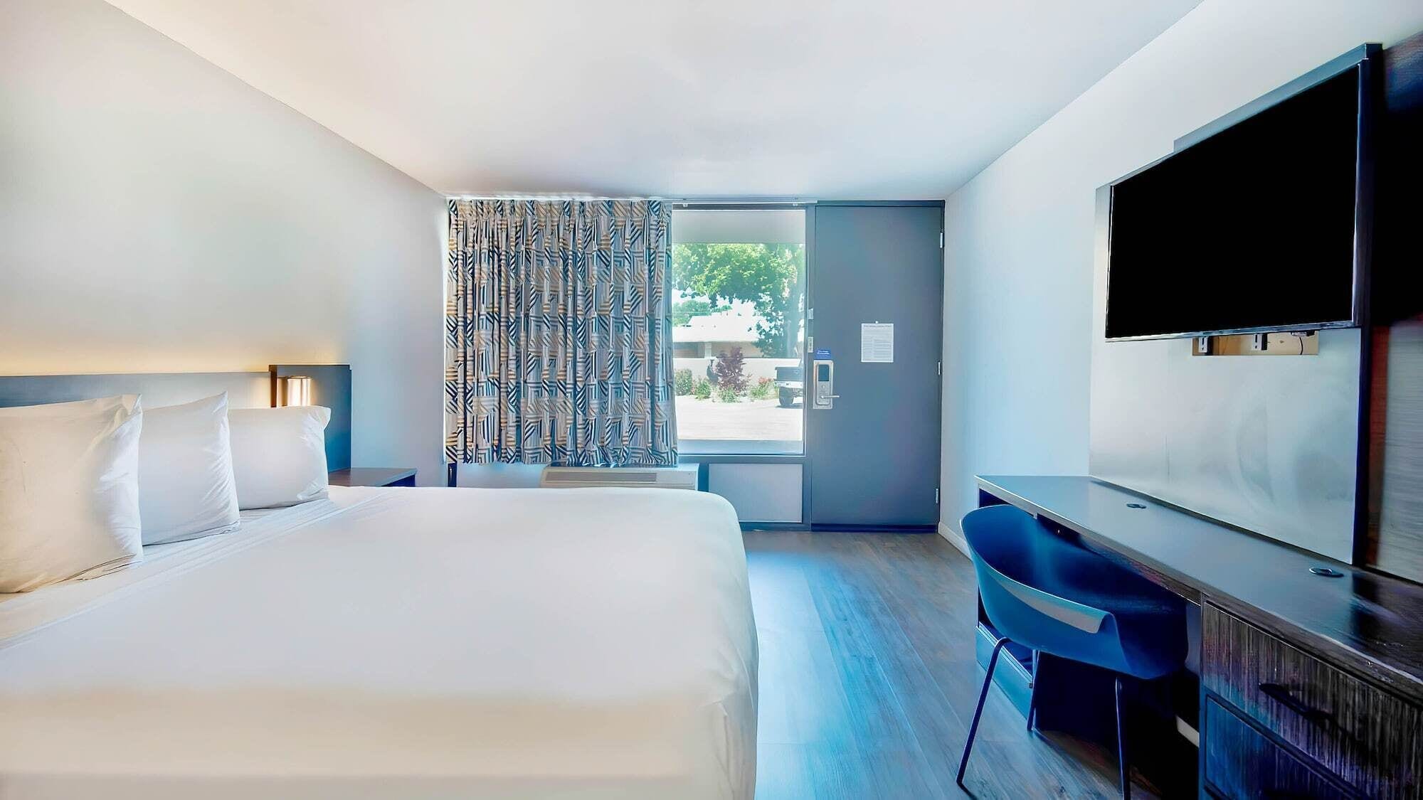A hotel room with a large bed, a desk, a chair, a TV, and a window with patterned curtains.