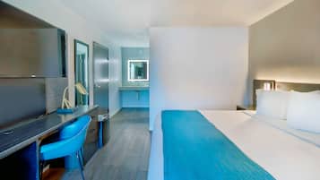 Camera d'albergo con un letto, una scrivania con una sedia, una TV, uno specchio e un bagno visibile attraverso una porta aperta.