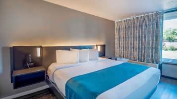 A hotel room with a large bed, a nightstand, a wall-mounted control panel, and a window with patterned curtains.