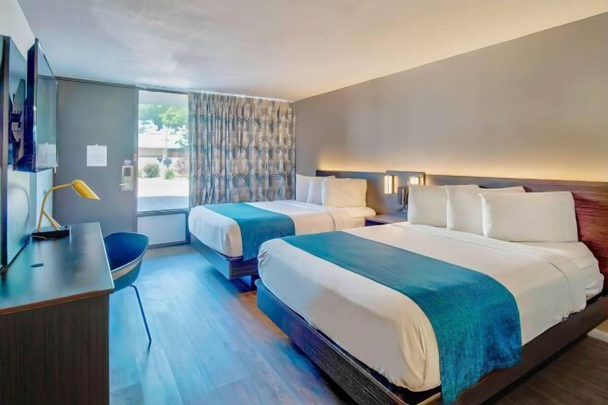 Ein Hotelzimmer mit zwei Betten, einem Schreibtisch, einem Stuhl und einem großen Fenster mit Vorhängen.