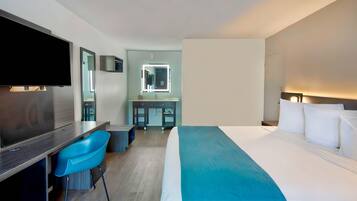 A hotel room with a bed, a desk with a chair, a TV, a mirror, a bathroom, and a nightstand with a lamp.