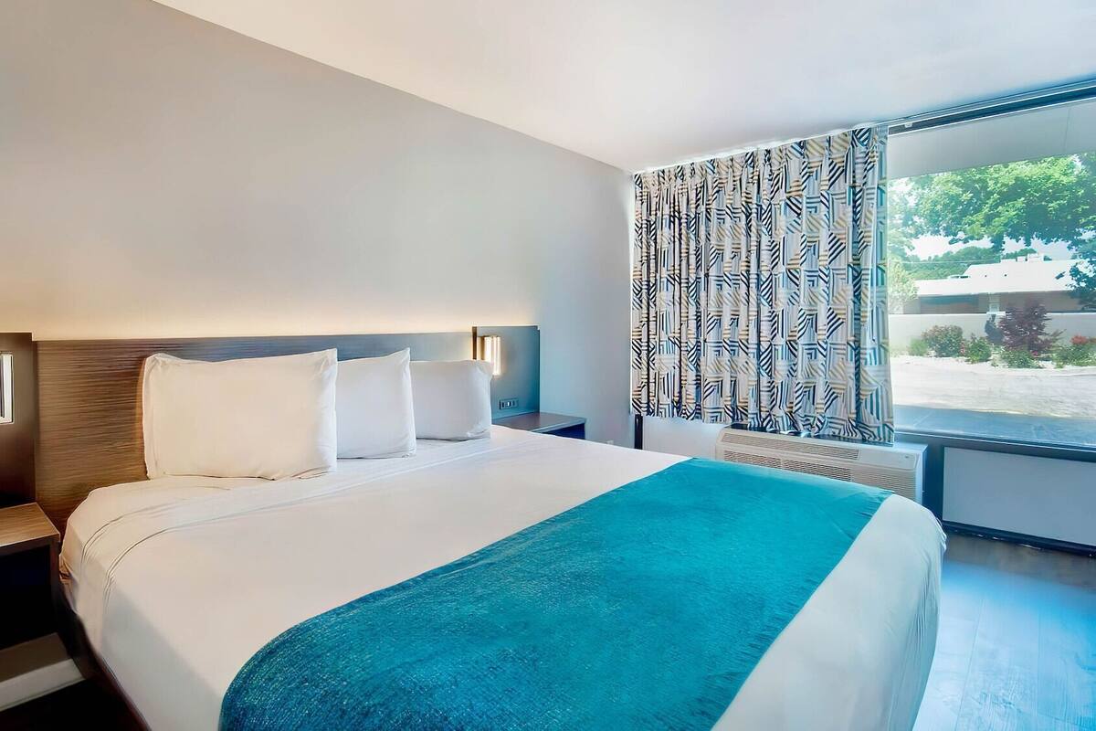 Ein modernes Hotelzimmer mit einem großen Bett, einem Nachttisch mit Lampe und Blick ins Freie durch einen gemusterten Vorhang.