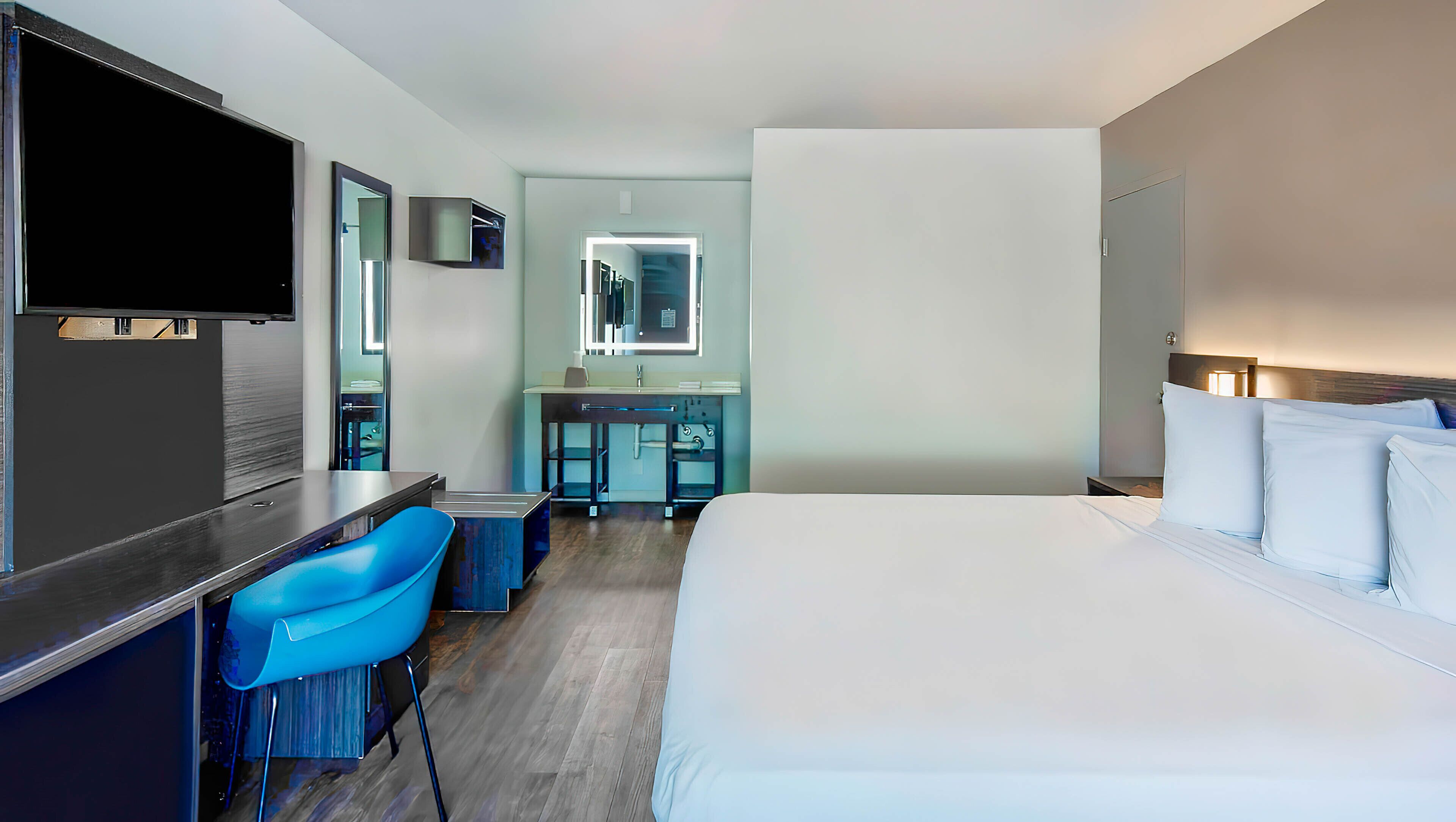 A hotel room with a bed, a desk with a chair, a TV, a mirror, and a bathroom with a sink and mirror.