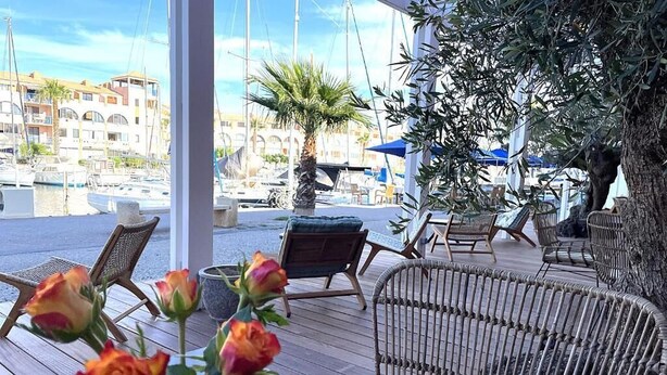 A covered outdoor seating area with wicker furniture and a view of the marina. - Hôtel des 2 golfs (Leucate)